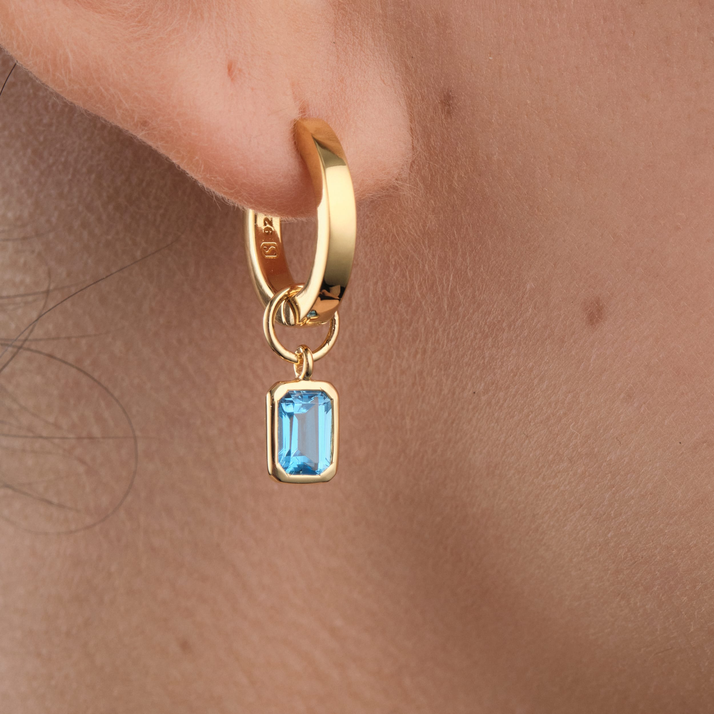 Close-up of 18K gold vermeil huggie hoop earrings with customisable blue topaz gemstone charms. A timeless and budget-friendly accessory showcasing December’s birthstone, crafted from durable gold vermeil for long-lasting wear.