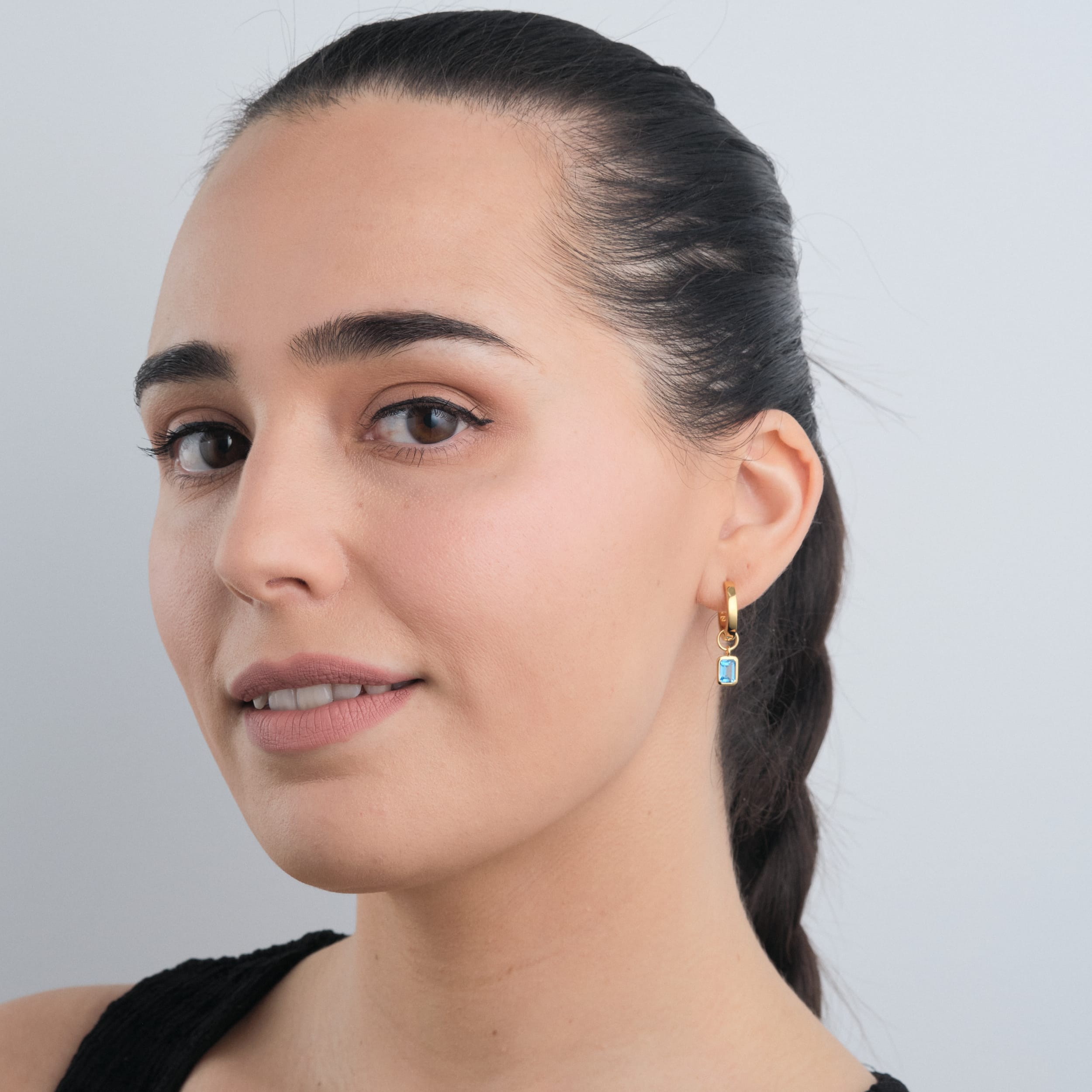 A woman wearing 18K gold vermeil huggie hoop earrings with interchangeable blue topaz gemstone charms. A versatile and elegant choice featuring December’s birthstone, crafted with long-lasting, high-quality gold vermeil.