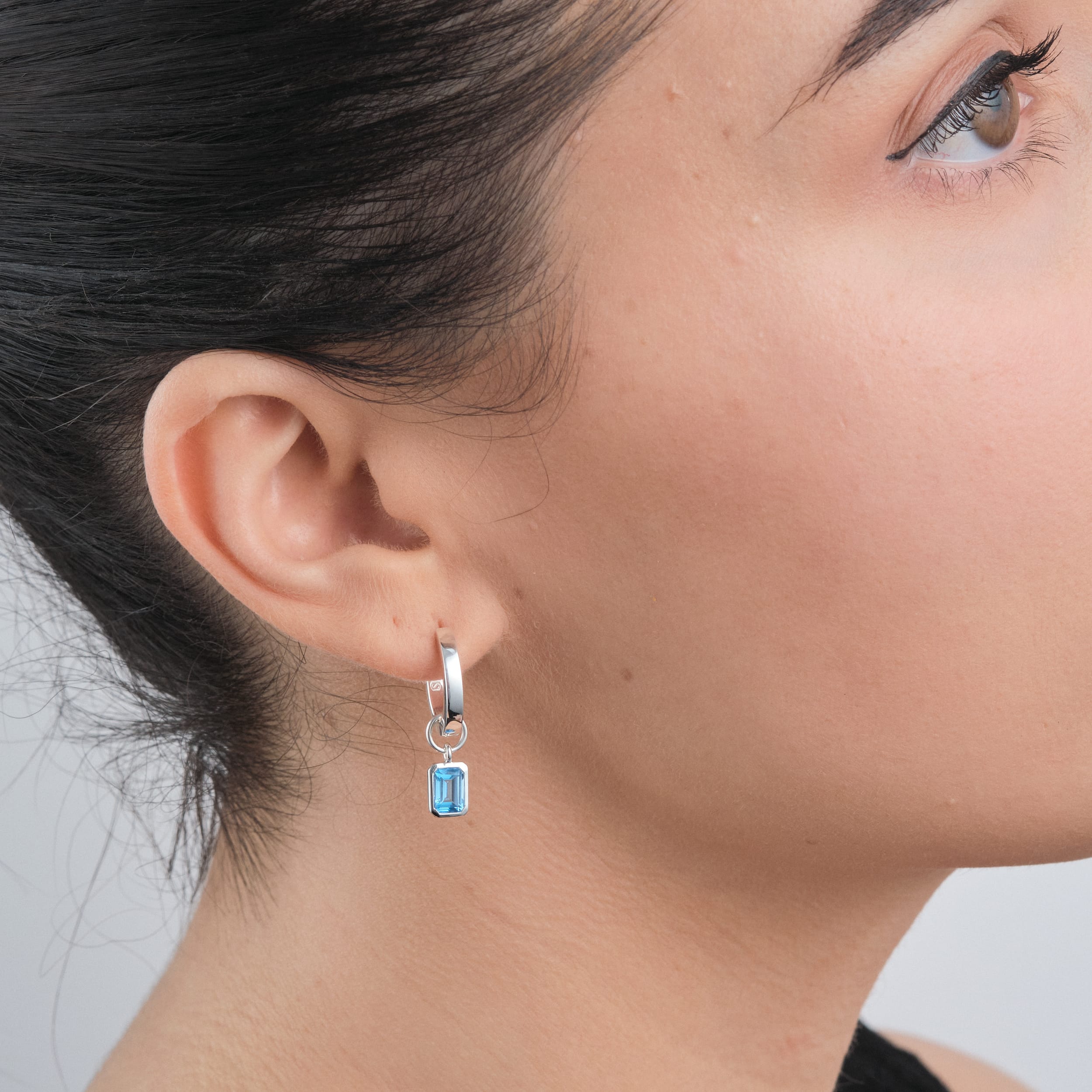 Side view of a woman wearing tarnish free sterling silver blue topaz charms attached to an elegant pair of huggie hoop earrings.