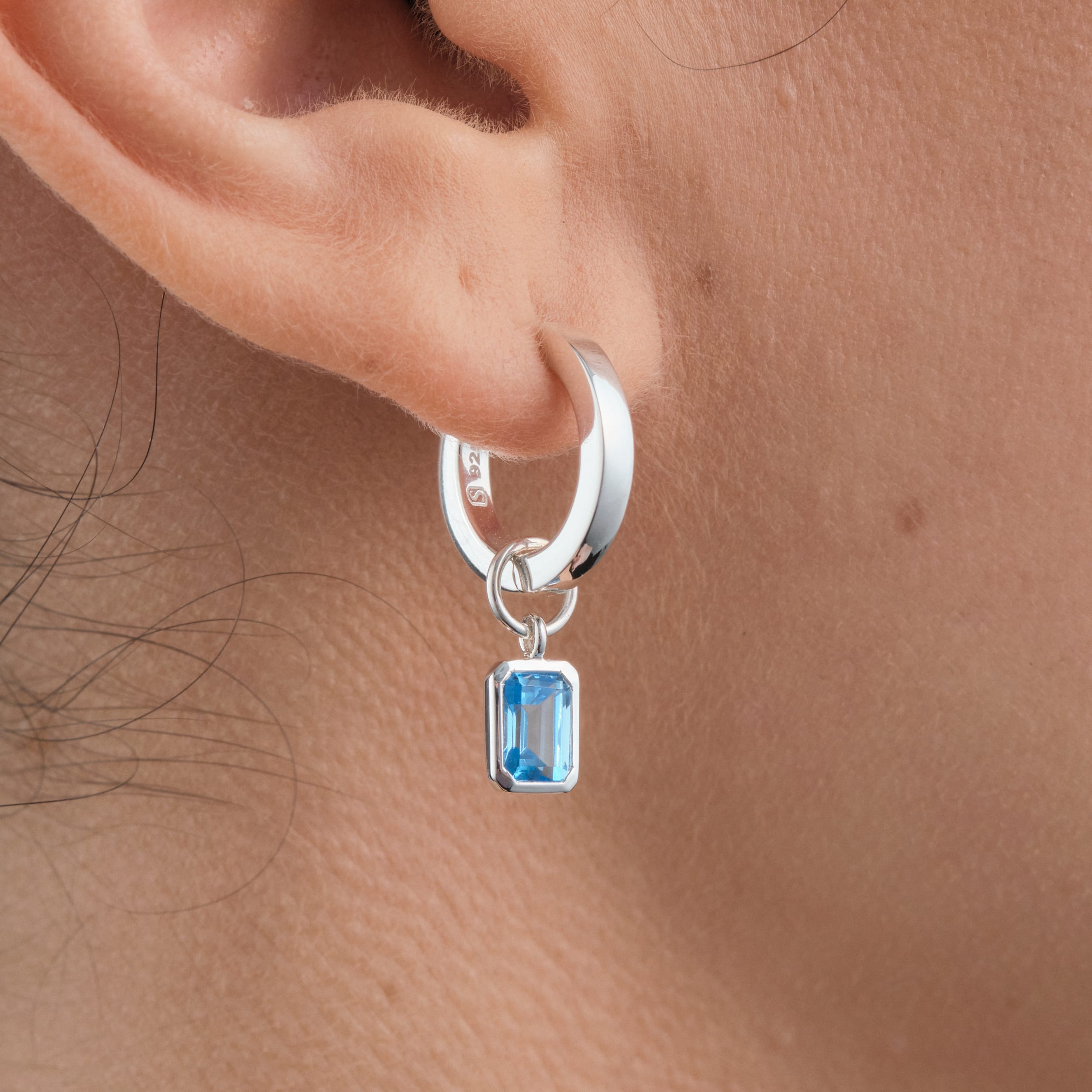 Close-up of sterling silver huggie hoop earrings with interchangeable blue topaz gemstone charms. Made with hypoallergenic sterling silver and a vibrant blue topaz make these earrings the perfect customisable piece for any occasion. The perfect December birthstone gift.