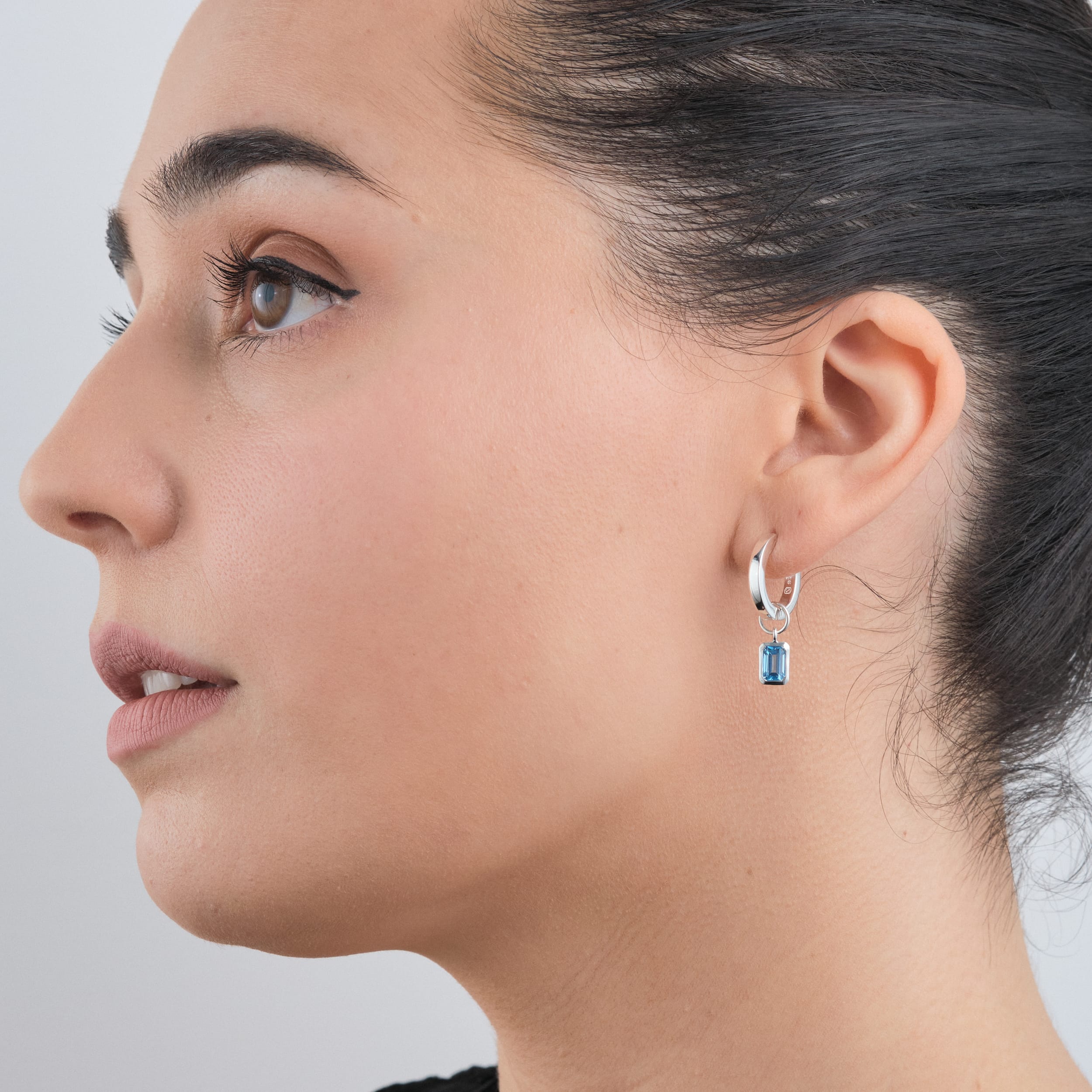 Side view of a woman wearing sterling silver huggie hoop earrings with interchangeable December birthstone blue topaz gemstone charms. Designed for comfort and versatility, these earrings are made from durable, hypoallergenic sterling silver, a perfect gift for those who love personalised jewellery.