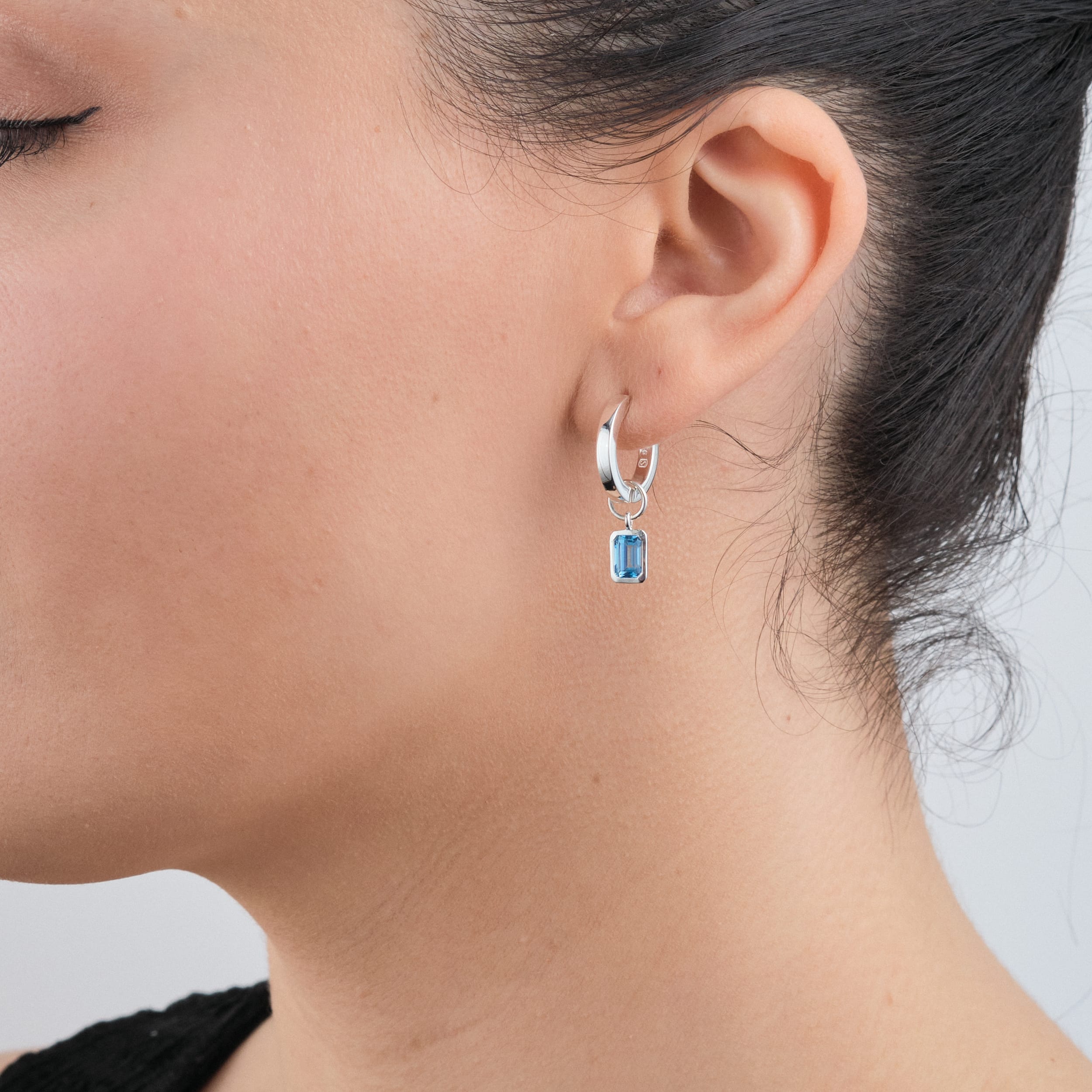 Side view close up of a woman wearing Sterling silver huggie hoop earrings with interchangeable blue topaz gemstone charms. Crafted from durable, hypoallergenic sterling silver, these earrings provide an affordable yet elegant gift option for birthdays in December, perfect for those who appreciate versatile, customisable jewellery.