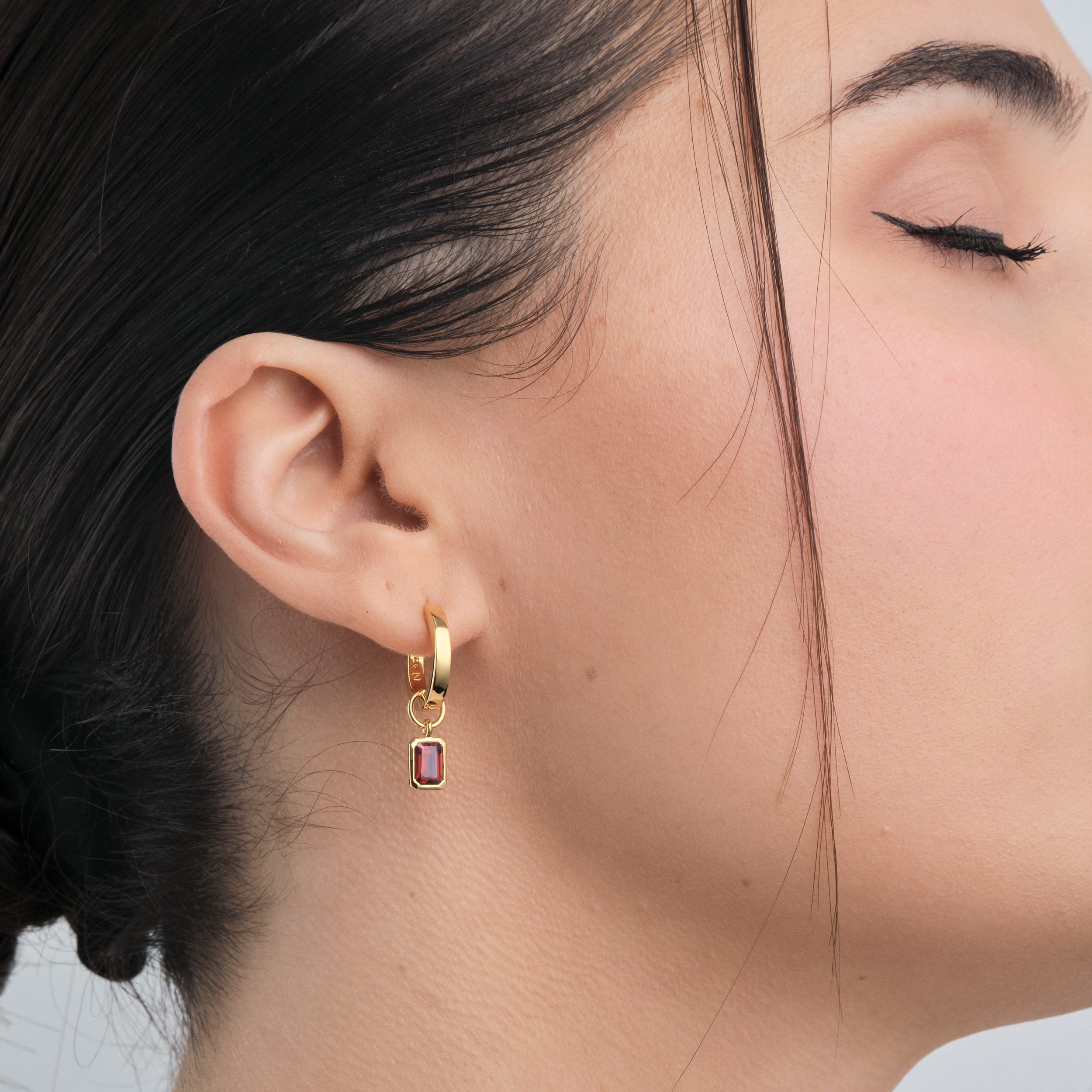 Side view of a woman wearing 18K gold vermeil huggie hoop earrings with interchangeable red garnet gemstone charms. An affordable yet sophisticated way to wear January’s birthstone, with the lasting quality and shine of gold vermeil.