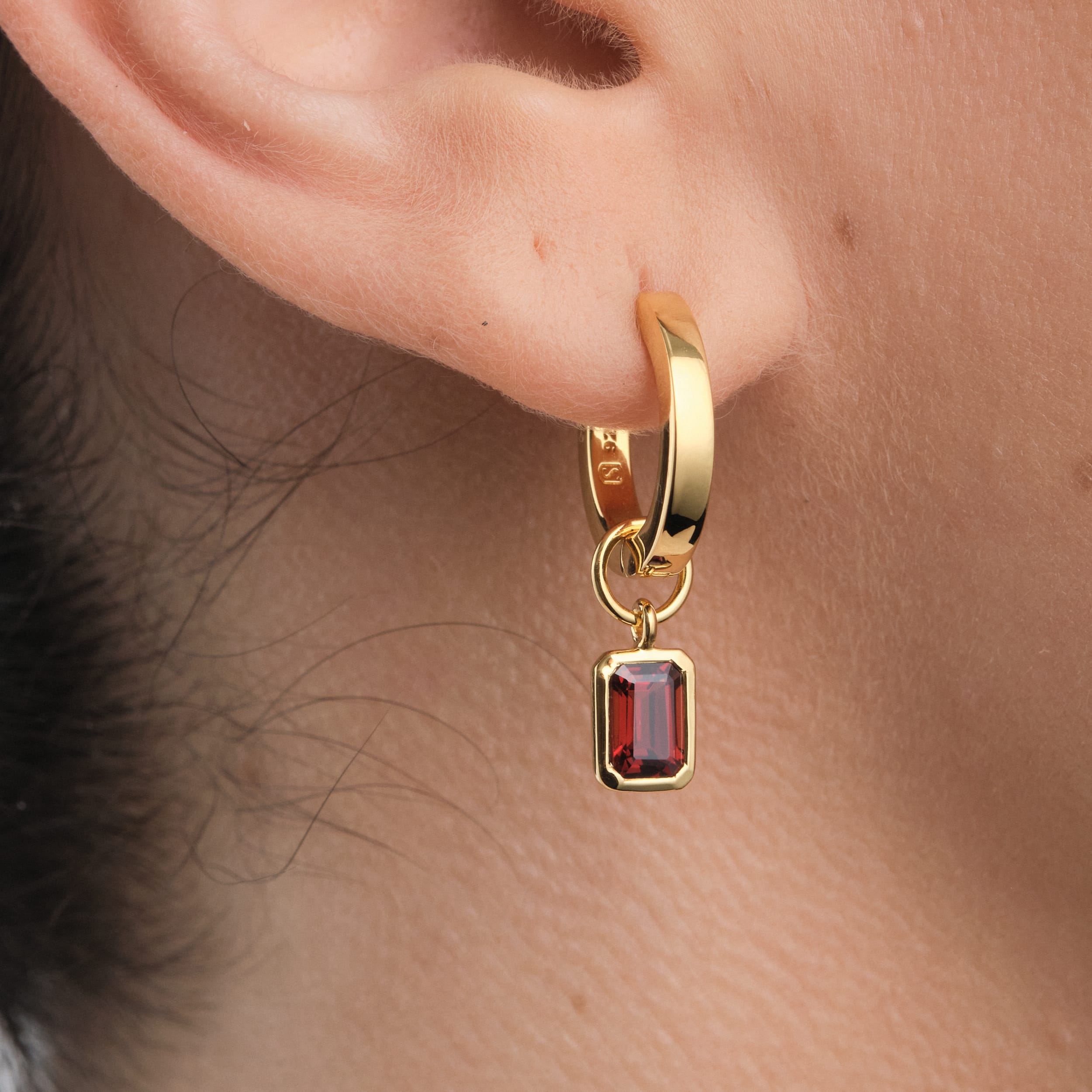 Close-up of a woman wearing 18K gold vermeil huggie hoop earrings with interchangeable red garnet gemstone charms. A luxurious yet budget-friendly jewellery piece, combining the bold beauty of January’s birthstone with the long-lasting quality of gold vermeil.