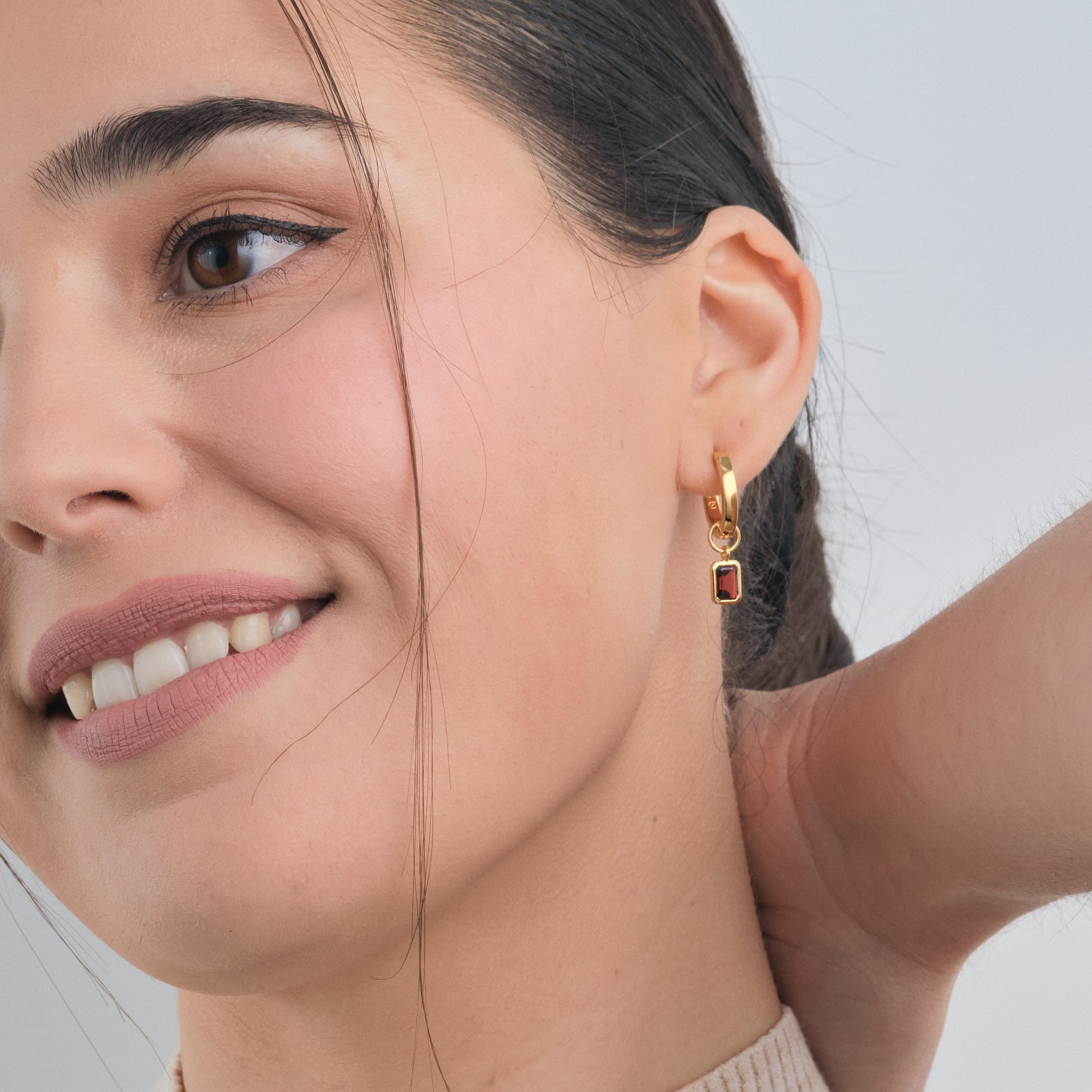 Woman wearing 18K gold vermeil huggie hoop earrings with customisable red garnet gemstone charms – designed for those who love meaningful jewellery, these durable and affordable earrings showcase January’s birthstone in a versatile, elegant setting.