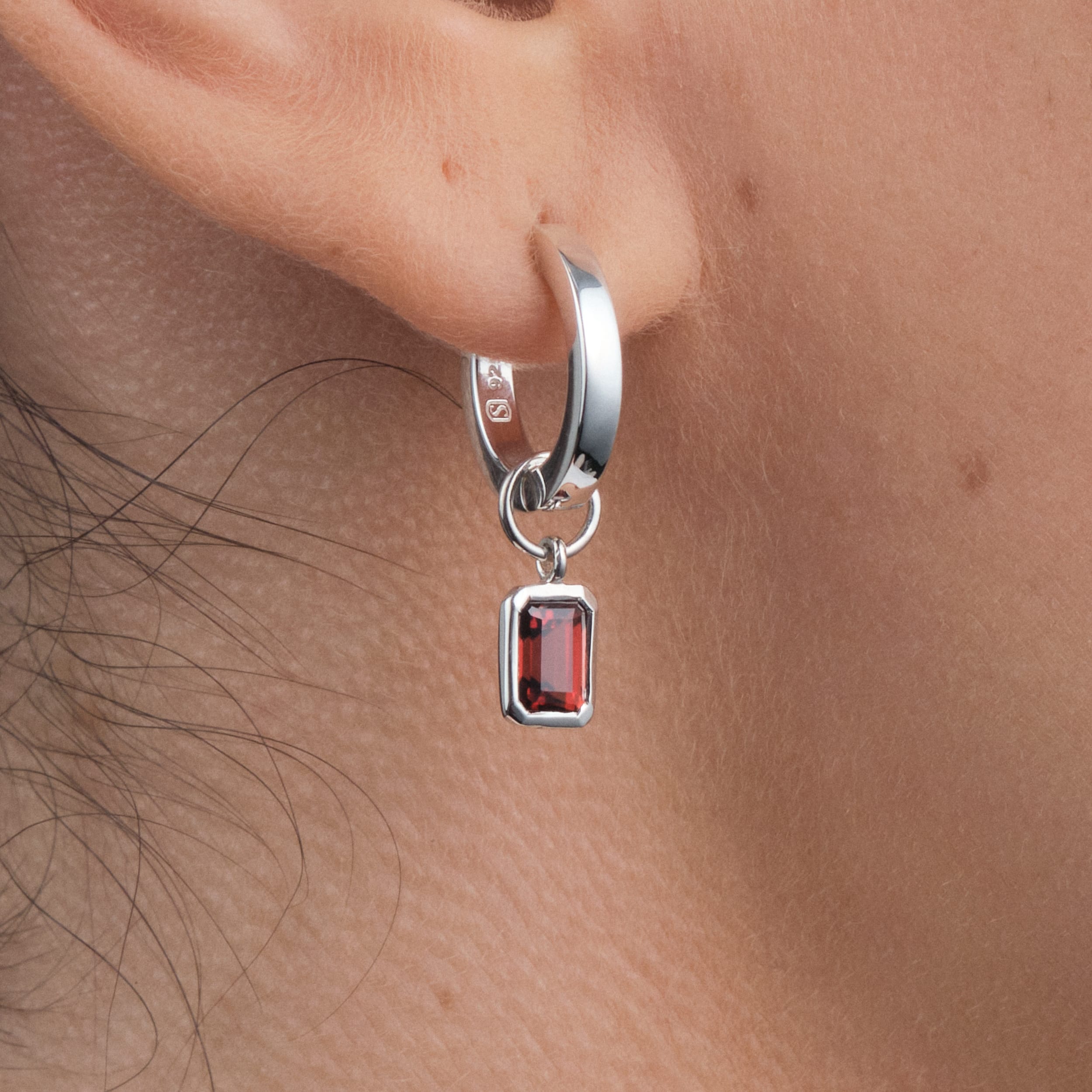 Close-up of sterling silver huggie hoop earrings with interchangeable red garnet gemstone charms – durable, hypoallergenic sterling silver paired with red garnet, the January birthstone, makes these earrings a versatile, customisable luxury piece.