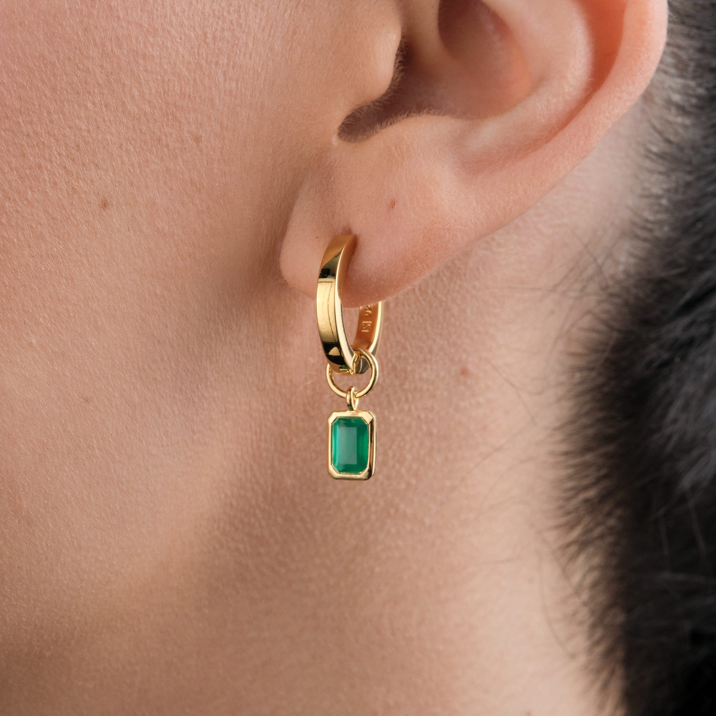 Side front view of a woman wearing 18K gold vermeil huggie hoop earrings with interchangeable green onyx gemstone charms. A bold and sophisticated addition to any collection, featuring May’s birthstone with the strength and affordability of durable gold vermeil.