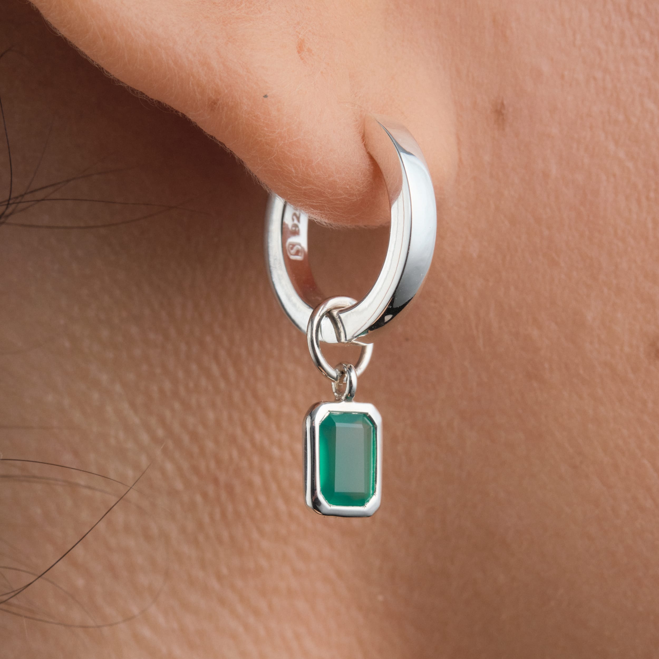 Close-up of sterling silver huggie hoop earrings with interchangeable green onyx gemstone charms. Durable, hypoallergenic sterling silver paired with green onyx, May's birthstone, makes these earrings a versatile and affordable luxury option.