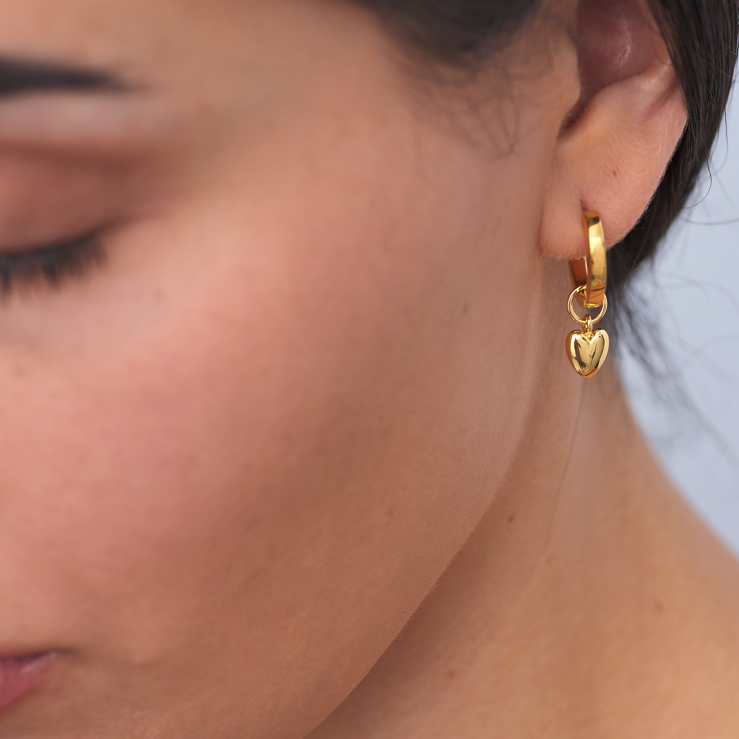 Close-up of a woman wearing 18K gold vermeil puffy heart charm huggie hoop earrings featuring interchangeable charms, allowing for versatile styling, a budget-friendly luxury gift for special occasions.