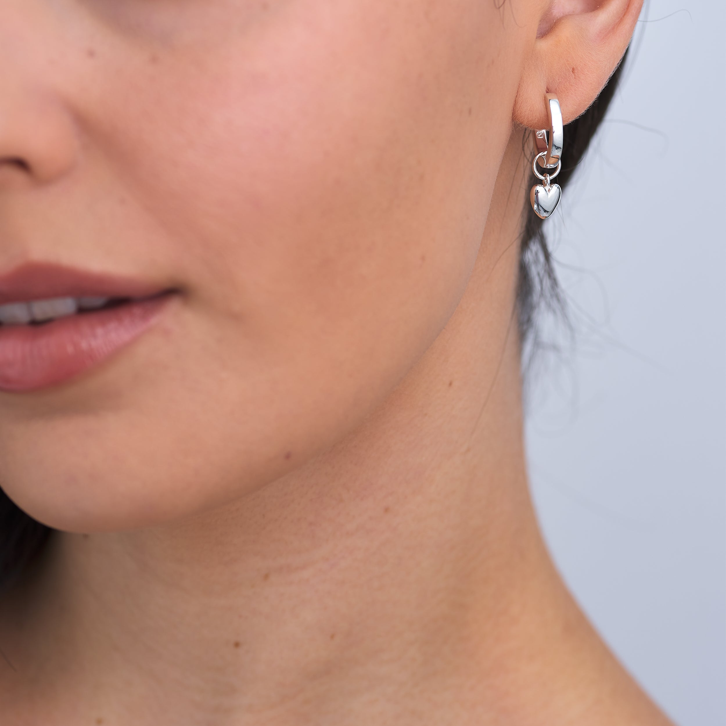 Close-up of a woman wearing puffy heart charm huggie hoop earrings in sterling silver, a timeless and versatile accessory, crafted from premium sterling silver for long-lasting wear. The perfect gift of love.