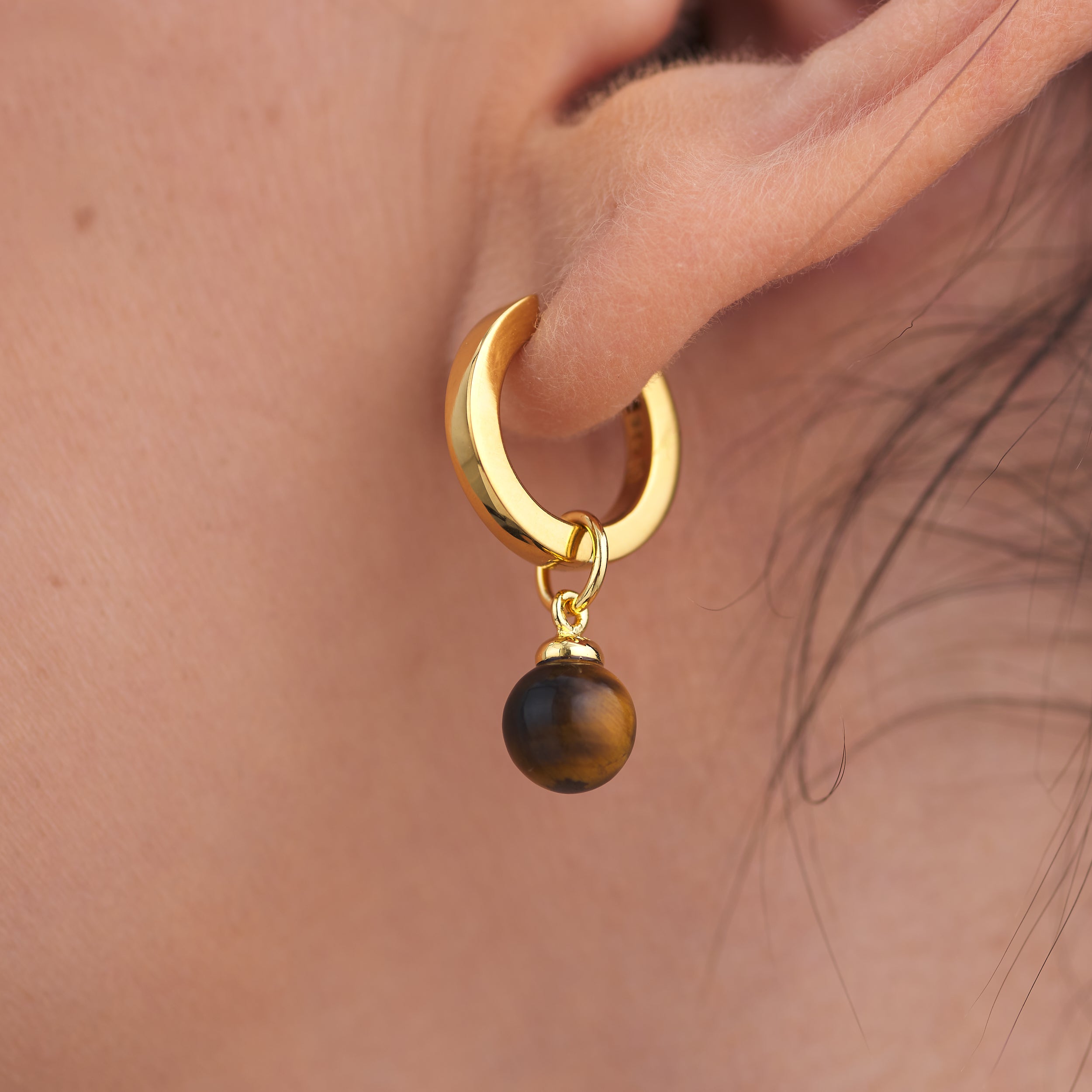 Luxurious 18k Gold Vermeil Huggie Hoop earrings with interchangeable Tiger's eye gemstone charm attached to waomn's earlobe.