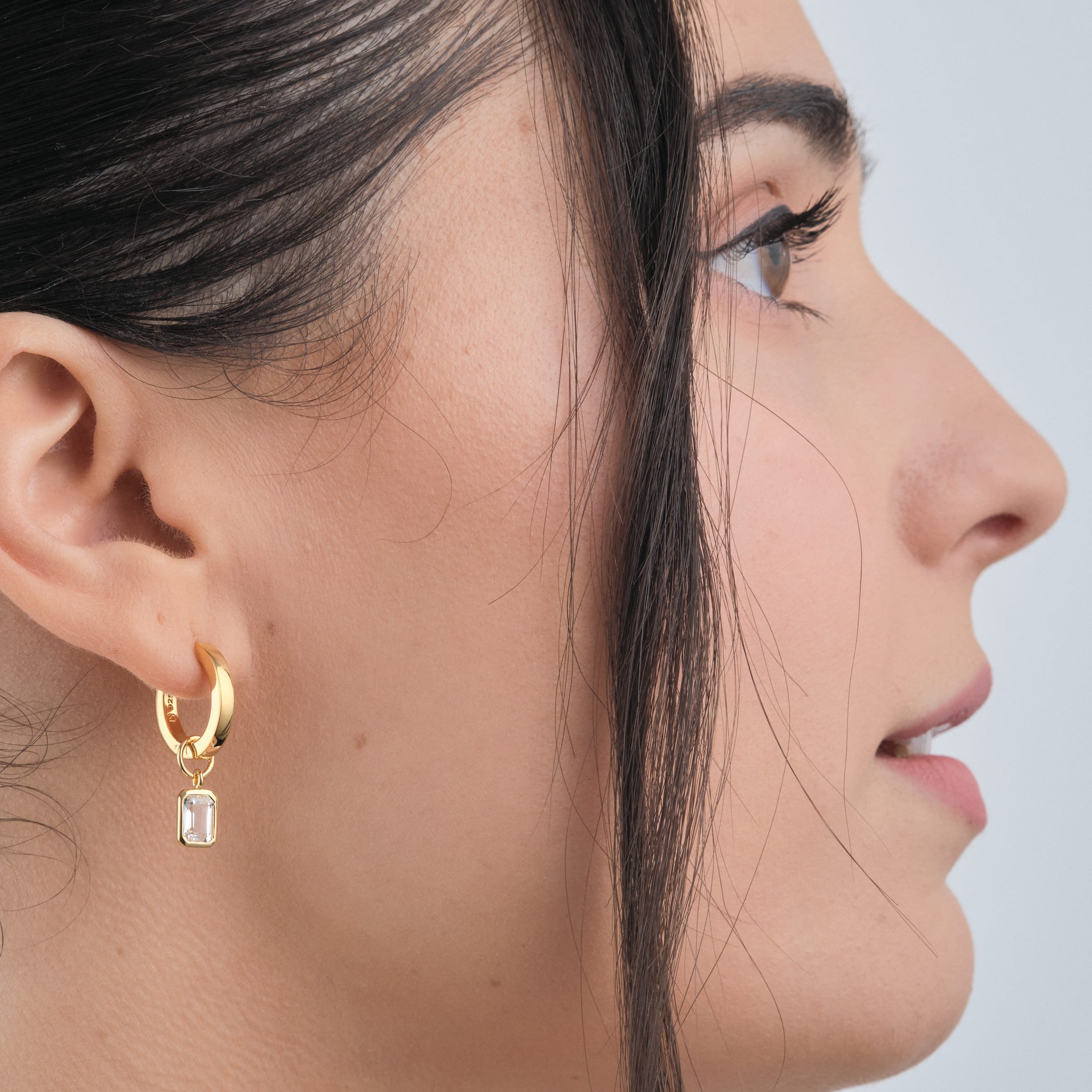 Side view of a woman wearing 18K gold vermeil huggie hoop earrings with interchangeable white topaz gemstone charms. Customisable and timeless, these durable and budget-friendly earrings are perfect for those who love elegant yet practical jewellery.