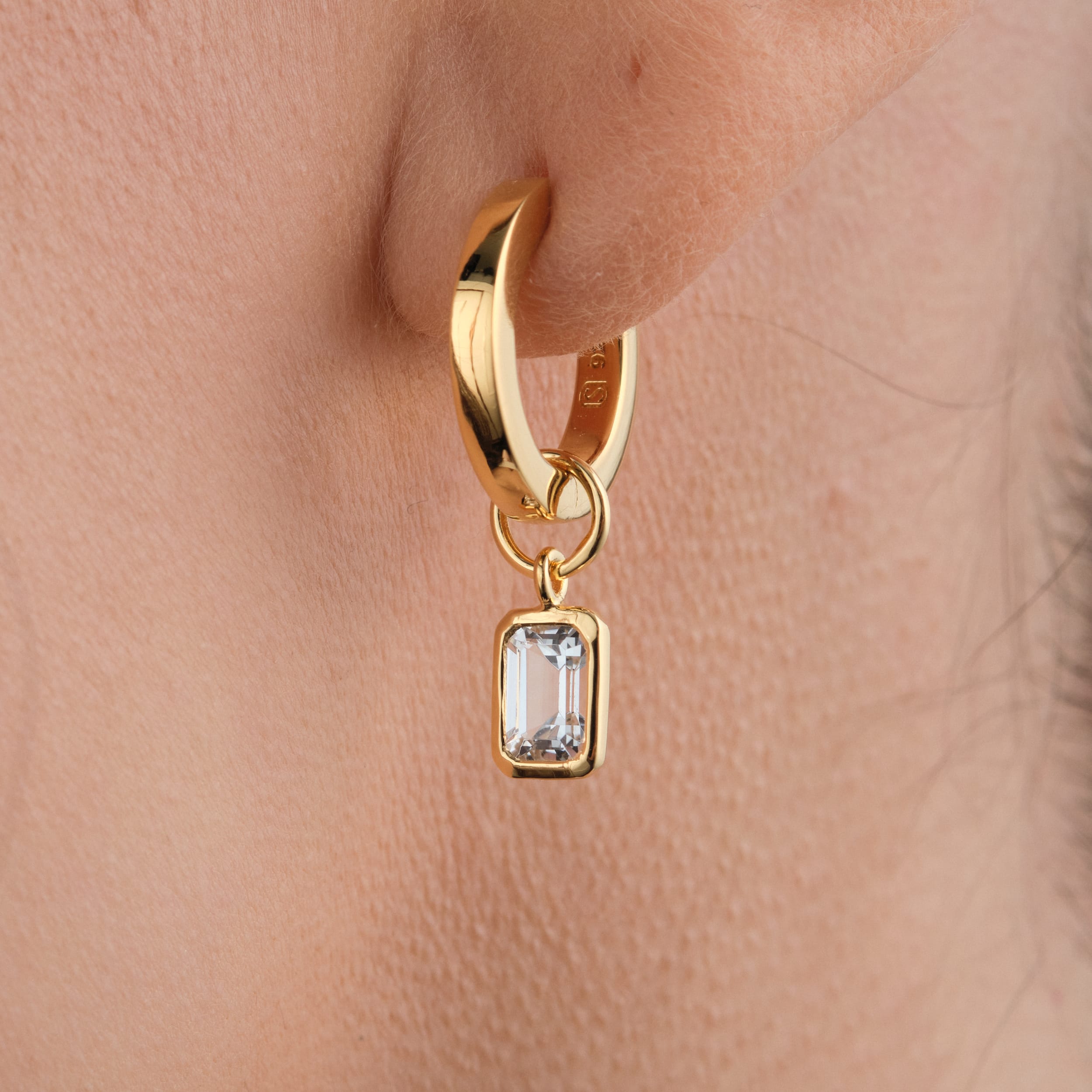 Close-up of a woman wearing 18K gold vermeil huggie hoop earrings with interchangeable white topaz gemstone charms made with high-quality, long-lasting gold vermeil, these earrings offer a sophisticated and budget-friendly way to elevate any look.
