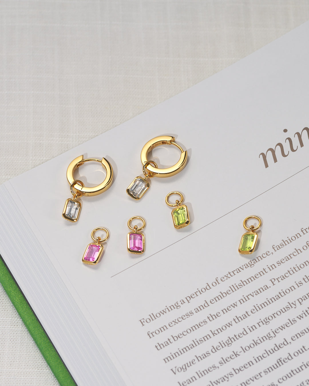 Four pairs of gold huggie hoop earrings with dangling rectangular gemstone charms in white topaz, pink ruby, peridot arranged on an open book page.