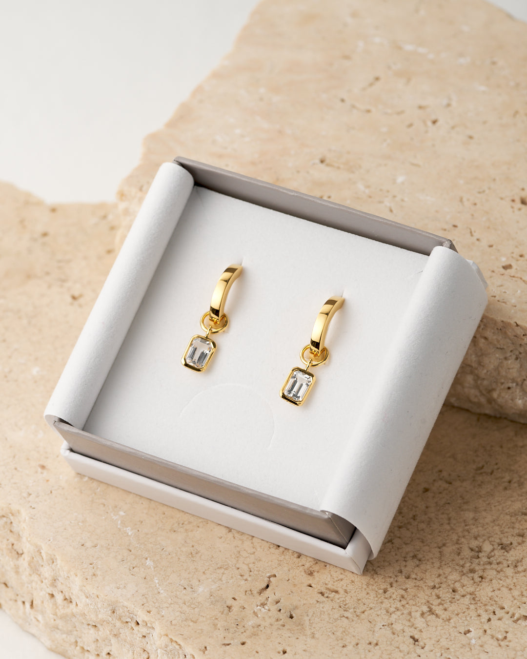 A pair of small gold huggie hoop earrings with rectangular white topaz stones, displayed inside an open white jewelry box on a beige stone surface.